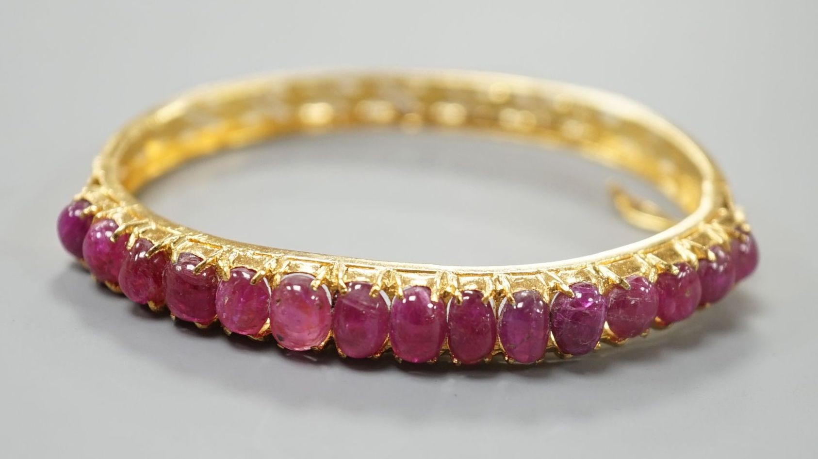 A modern Thai yellow metal and graduated fifteen stone cabochon ruby set hinged bangle, interior diameter 54mm, gross weight 12.9 grams.